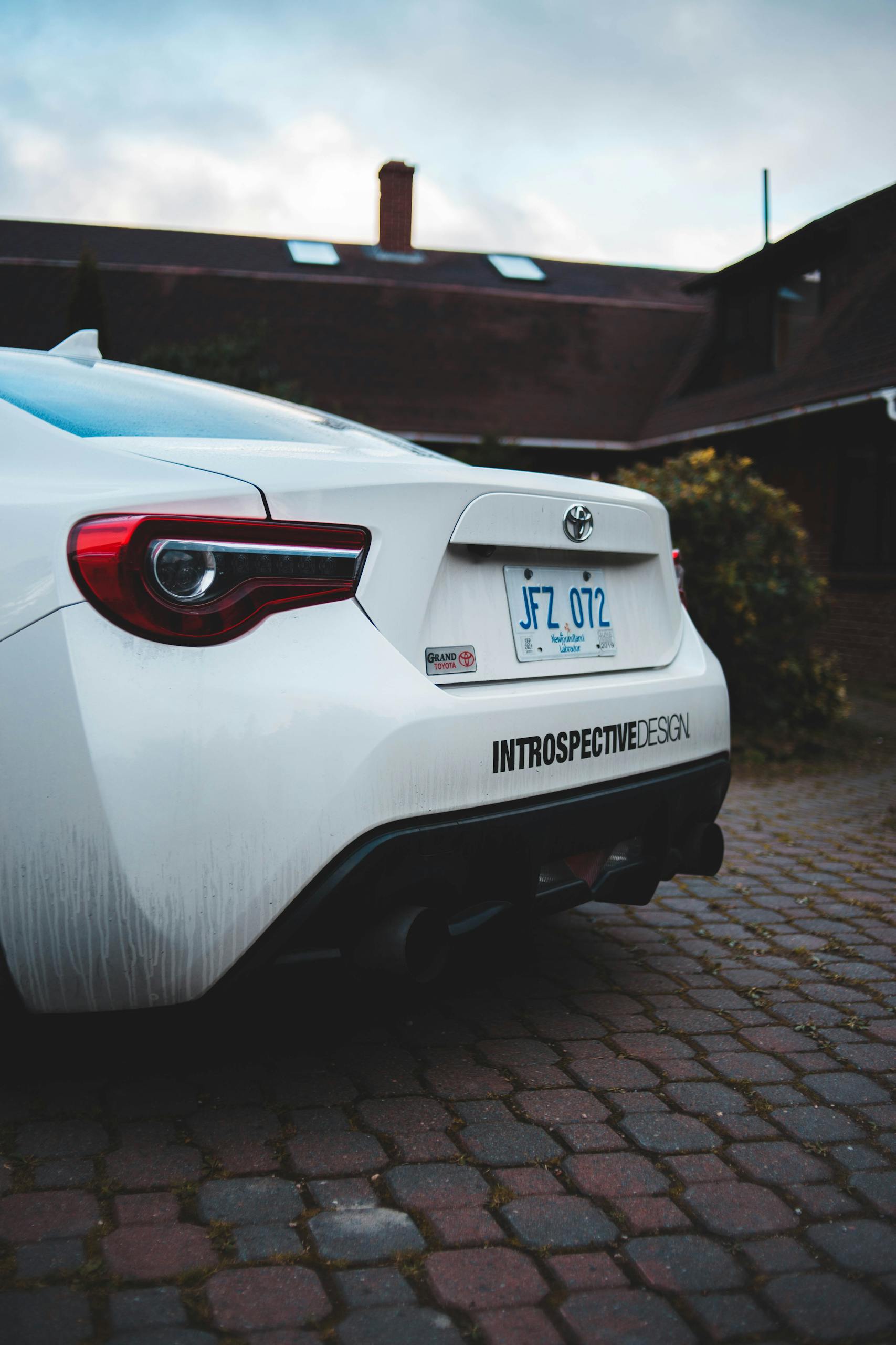 Close-up of a parked white sports car showing taillight and license plate on cobblestone driveway.