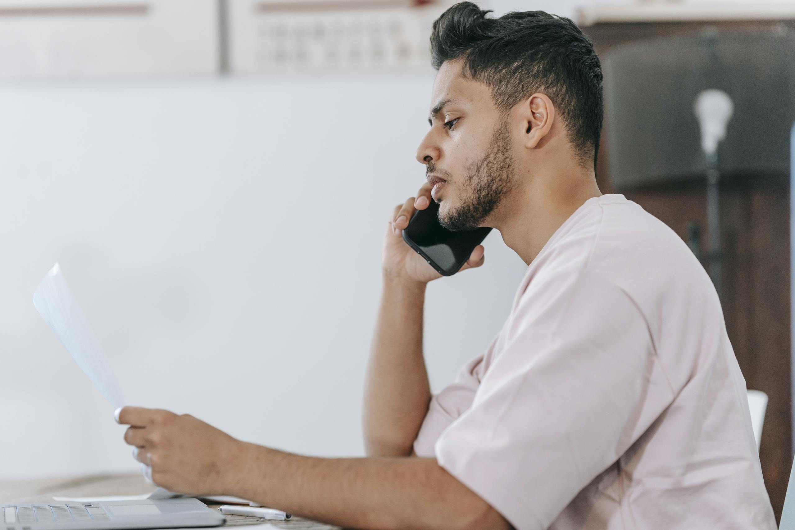 Side view thoughtful ethnic male in casual shirt reading document and talking on mobile phone while sitting at desk